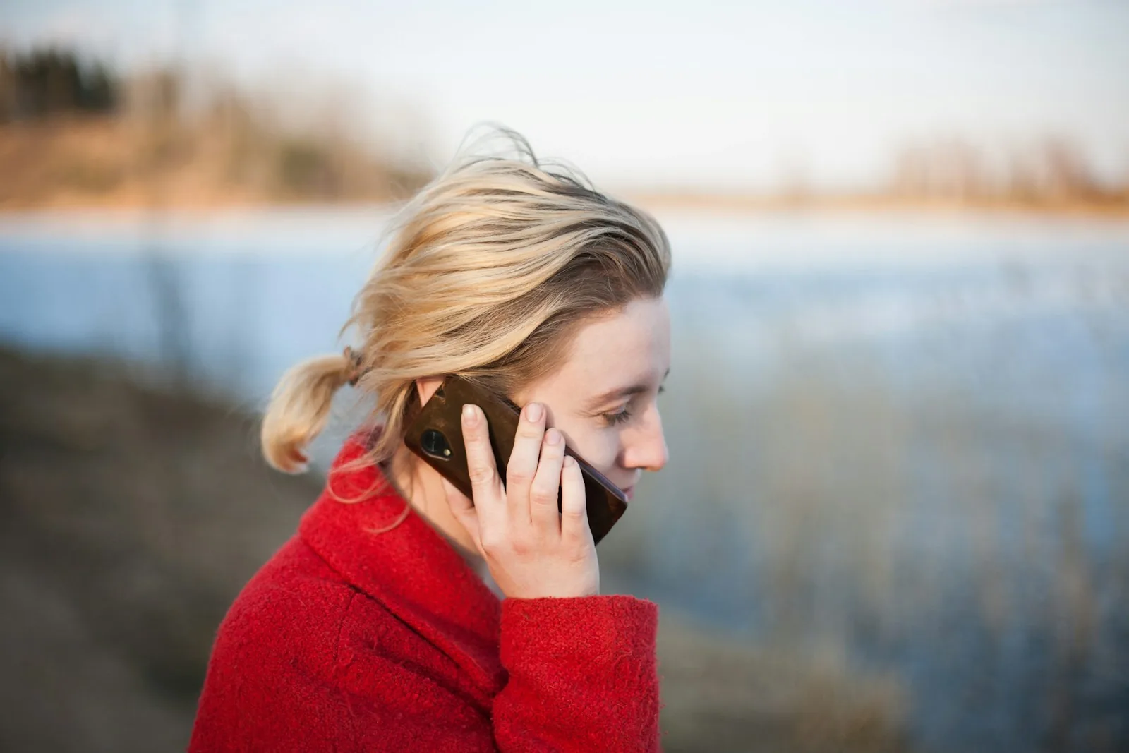 A woman in a red coat holding a smartphone to her ear outdoors beside a lake