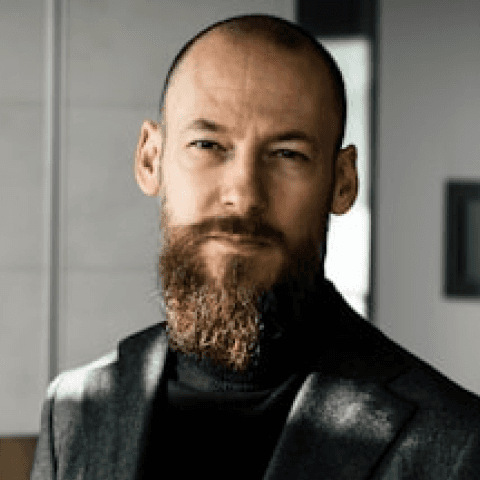 A circular headshot of a man with a shaved head and a full, well-groomed brown beard