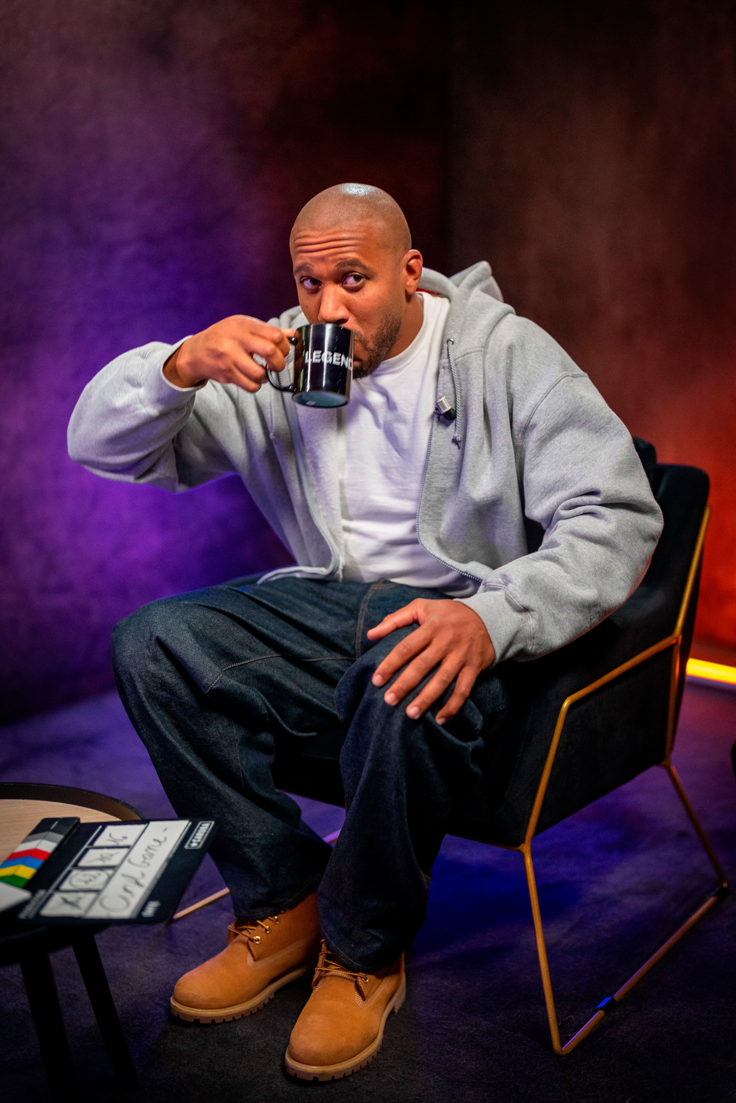 Photographie professionnelle du combattant de MMA français, Cyril Gane, en pleine interview pour Legend. Cyril Gane est assis sur un fauteuil moderne, buvant dans une tasse noire 'LEGEND'. Il est habillé de façon décontractée (sweat à capuche gris, t-shirt blanc, jean sombre, bottes camel). La lumière est tamisée avec des tons d'ambiance violets et rouges, typiques d'un studio d'interview. Cette photo de sport/lifestyle, optimisée par retouche, illustre l'expertise de Nux-Studio en production photo, direction artistique et création de contenu de marque pour des personnalités du monde du sport.