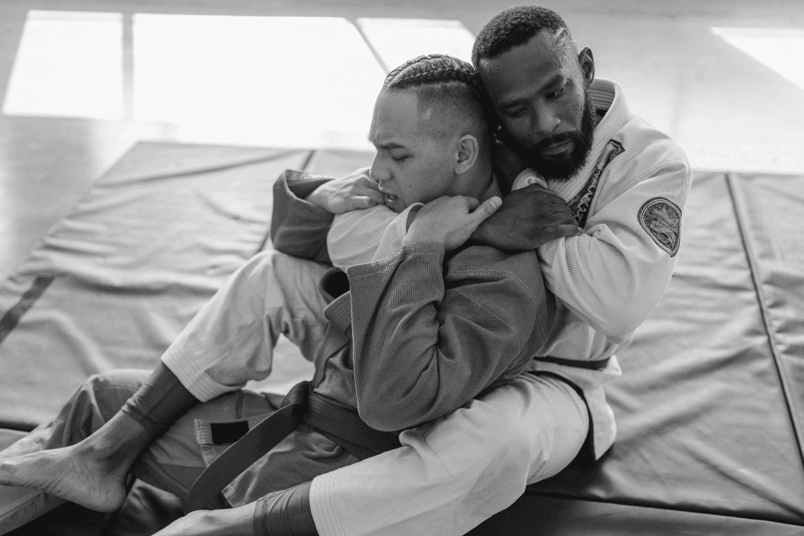 Close-up of two Jiu-Jitsu practitioners in gis, with one athlete in a white gi securing a back control hold on his training partner in a grey gi.