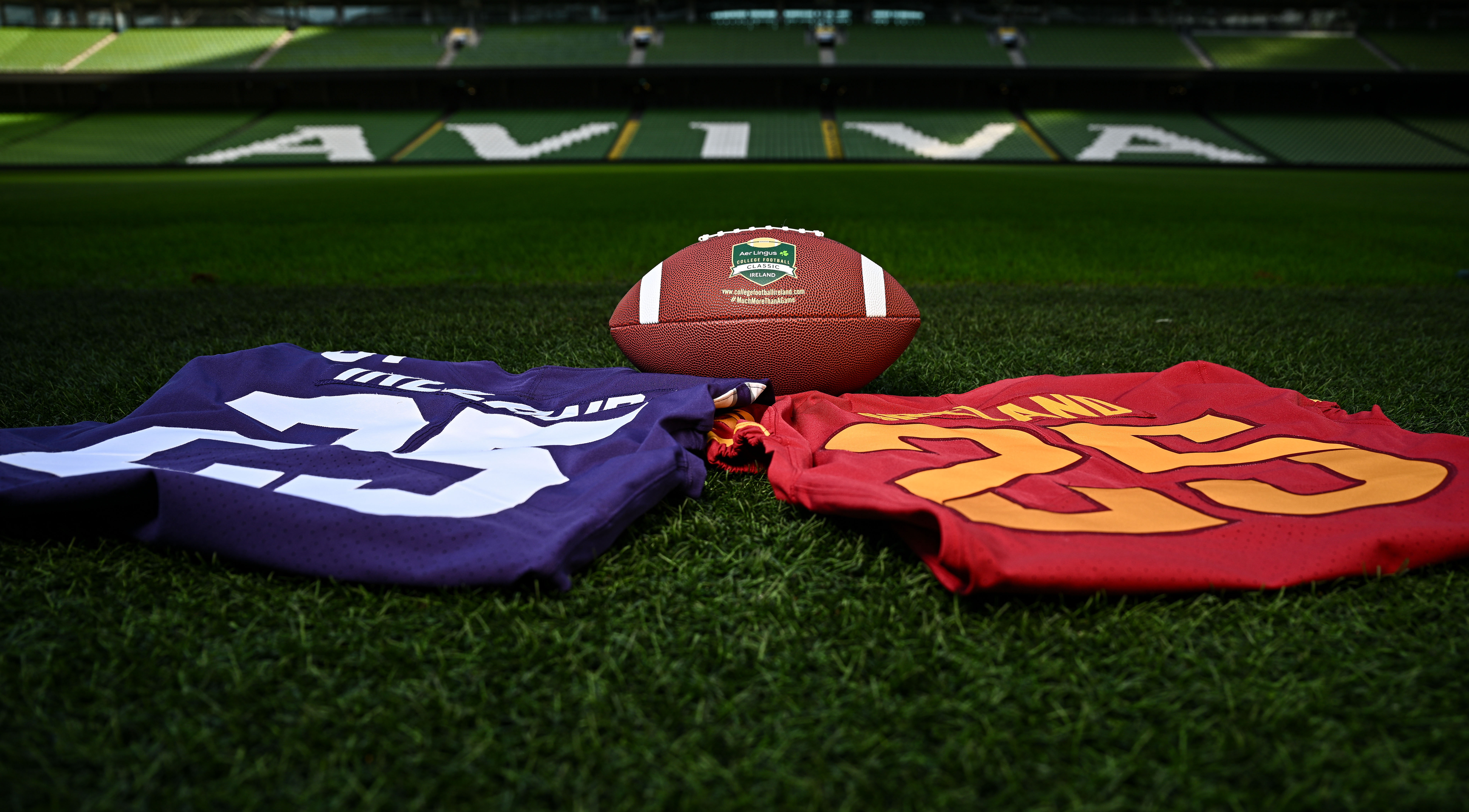 An American football with the Aer Lingus College Football Classic logo rests on the green turf alongside college jerseys in front of the Aviva Stadium. Investicon proudly partners with the "More Than Just a Game" initiative, connecting the worlds of investment and collegiate sports in Dublin. #Investicon #AerLingusCollegeClassic #CollegeFootball #DublinPartnership #SportsAndInvestment