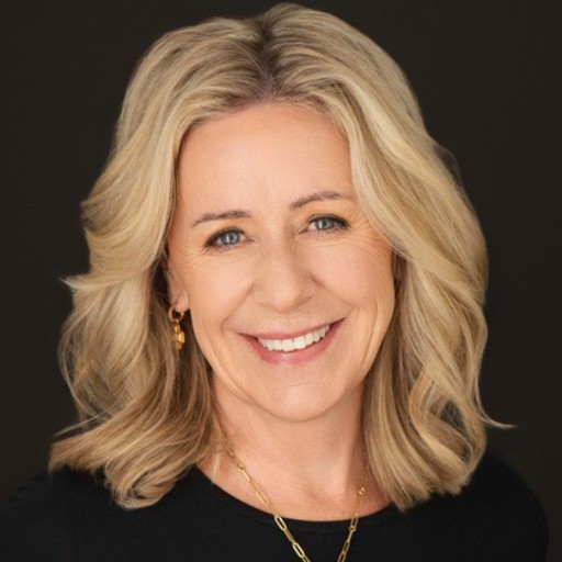 A focused woman with long red hair is seated at a desk, wearing a black blazer and dark blouse. She intently looks at camera, smiling slightly, with an open laptop in front of her. The setting is a warm and serene office setup in the background. Soft daylight filters through, creating a calm atmosphere. The composition captures her in a frontal profile with a clear emphasis on her concentration and the serene indoor setting.
