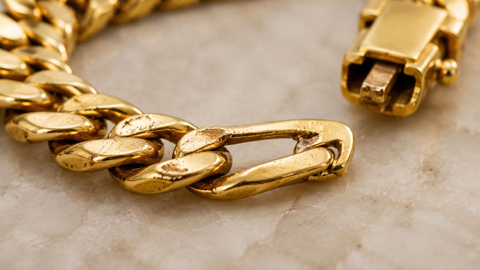 A close-up view of a luxury solid gold Cuban link bracelet resting on a textured marble surface.
