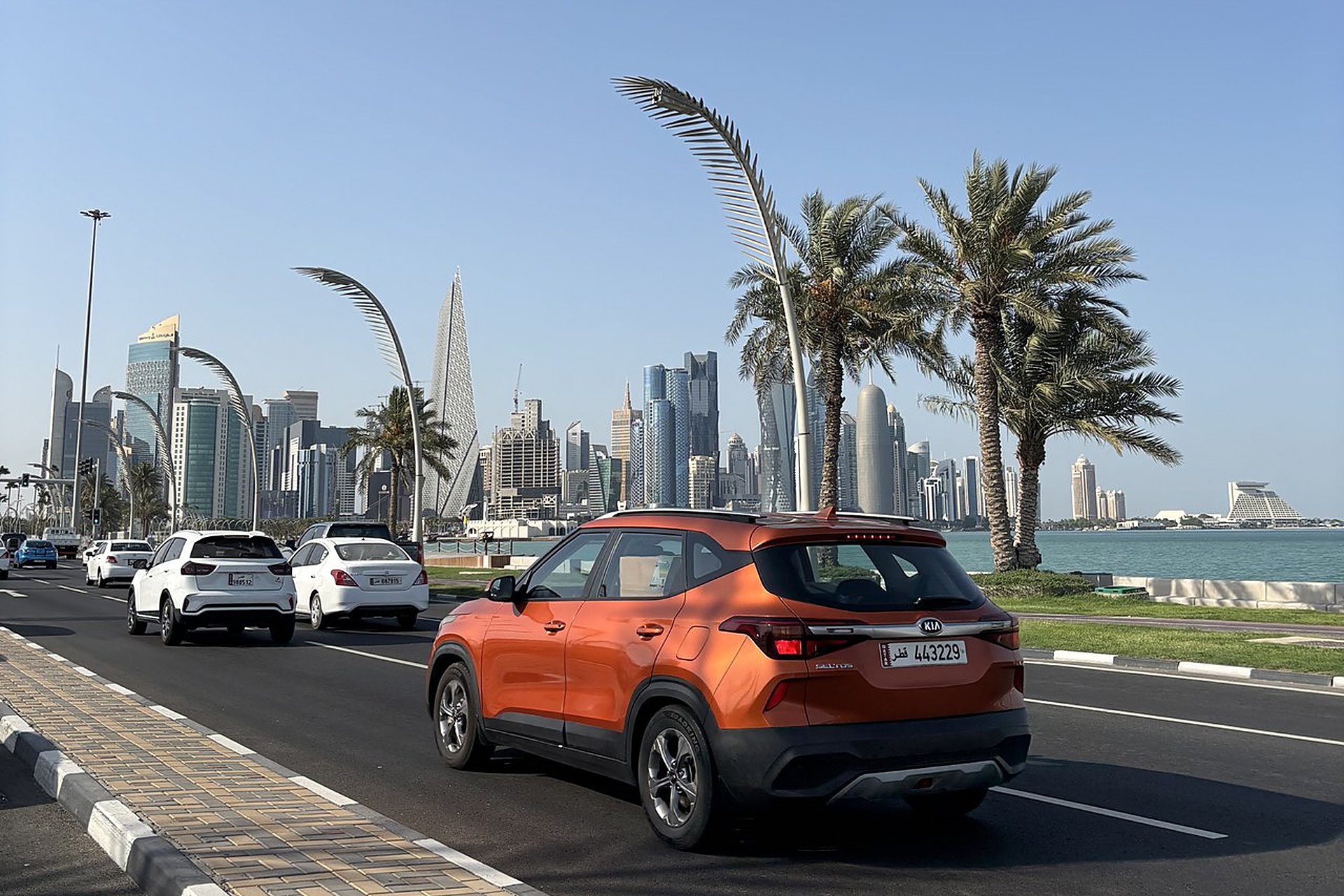 Une voiture orange sur la Corniche de Doha, palmiers et tours de West Bay au fond, une scène de tous les jours pour qui roule au Qatar