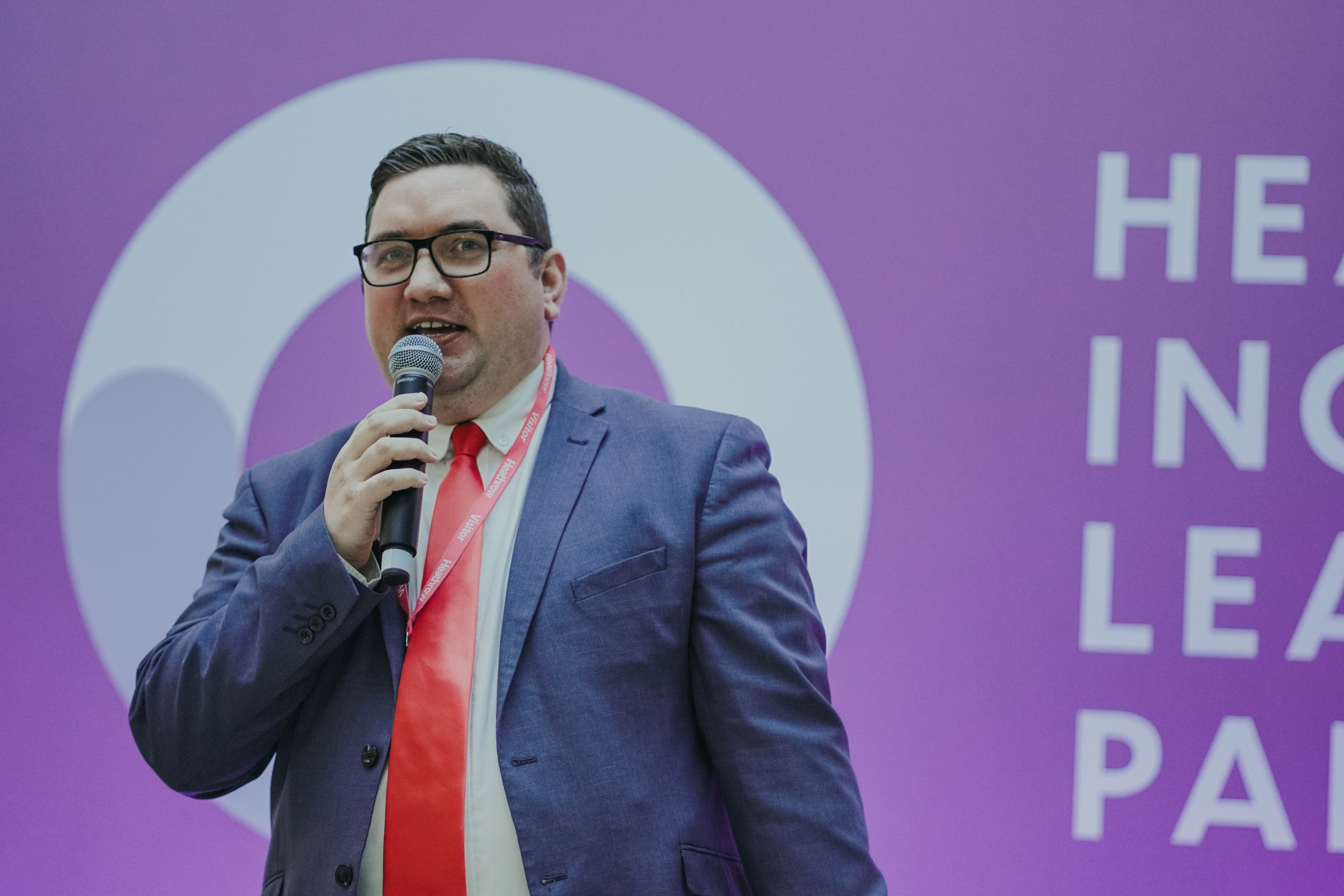 A man wearing glasses, a blue suit, and a red tie speaks enthusiastically into a microphone while standing in front of a large purple backdrop with white text.