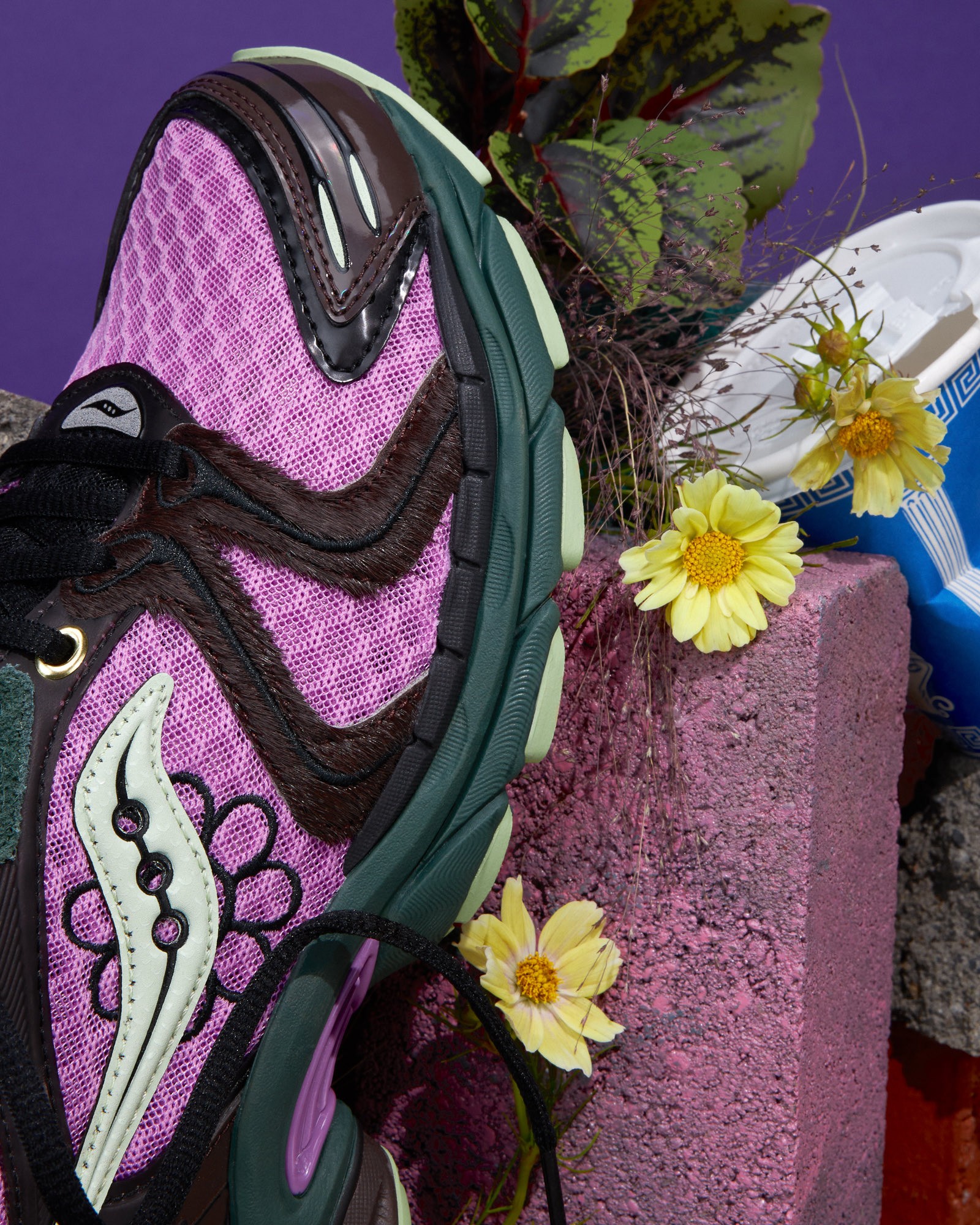 Purple Jae Tips x Saucony sneaker close-up on bricks with flowers and soil, showing layered textures and stitching.