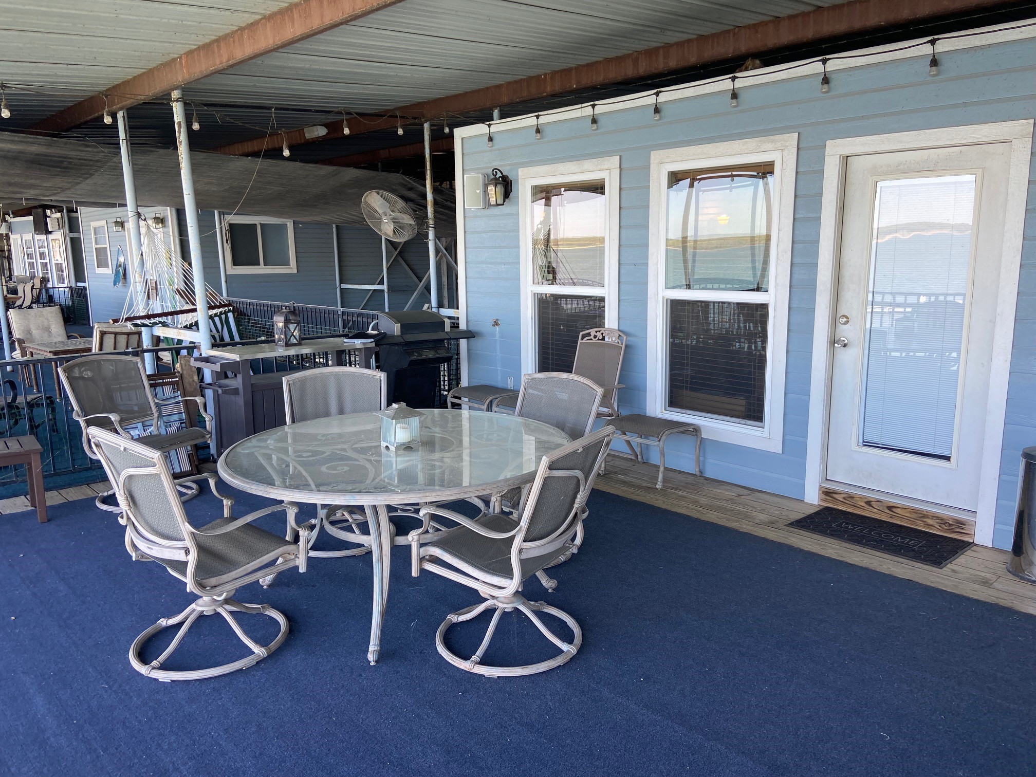 Outdoor patio with blue siding features a transparent circular glass table surrounded by six metal chairs on a dark blue carpet, set under a covered porch with string lights, adjacent to a built-in grill and closed white door with adjacent windows.
