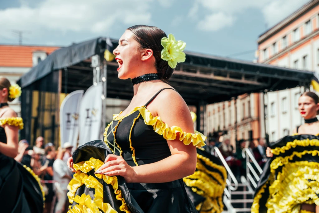 A woman in a bright yellow and black outfit dances joyfully at an outdoor event, with a crowd in the background.