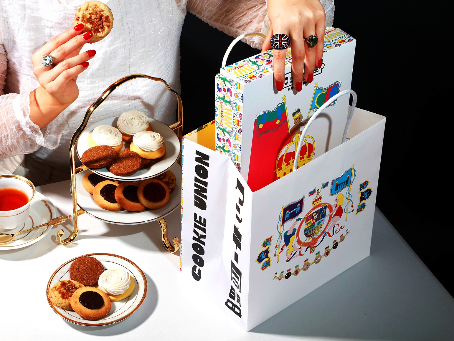 Cookie Union shopping bag and illustrated cookie box displayed beside a tiered plate of cookies, with a hand placing the box into the bag.