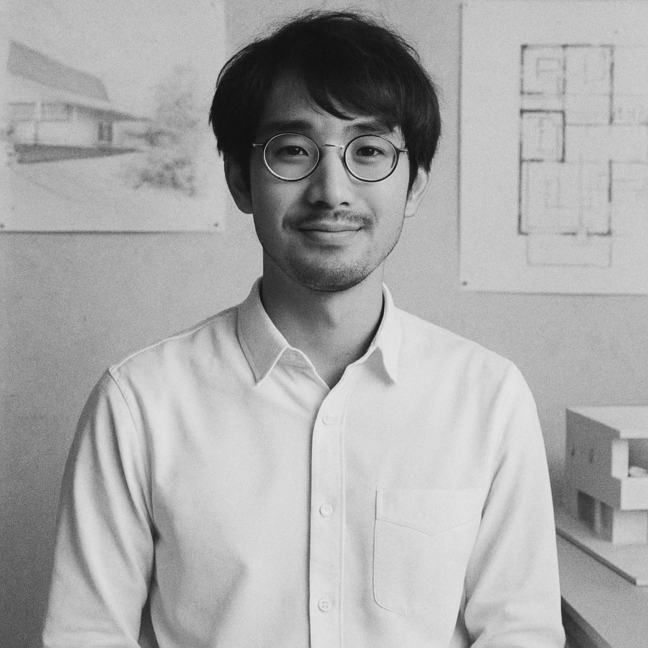 Black and white portrait of a young adult man with medium-length dark hair and round glasses, wearing a light-colored button-up shirt, standing in a room with architectural drawings visible on the wall behind him.