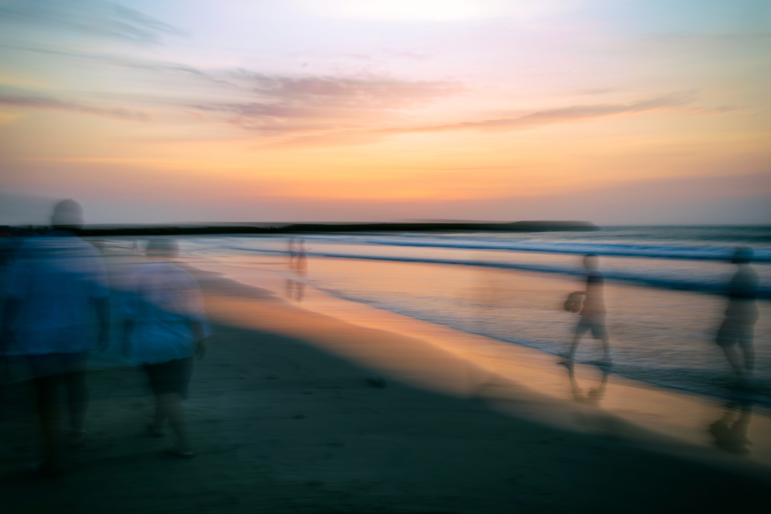 Ghost-like figures walking along the shoreline during a pastel sunset, captured with intentional blur