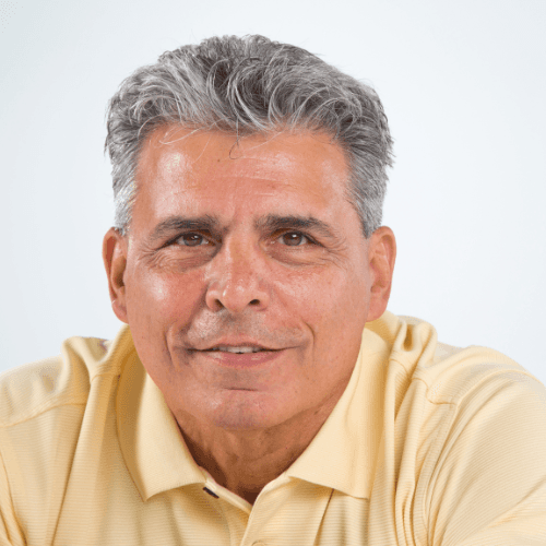 Portrait of a smiling man with curly hair and glasses, wearing a brown jacket over a dark shirt, standing against a neutral background.