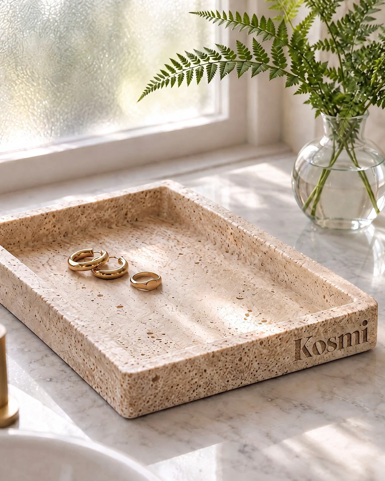 A travertine ring tray carved with the Kosmi logo, holding gold hoops and a ring, by a window.