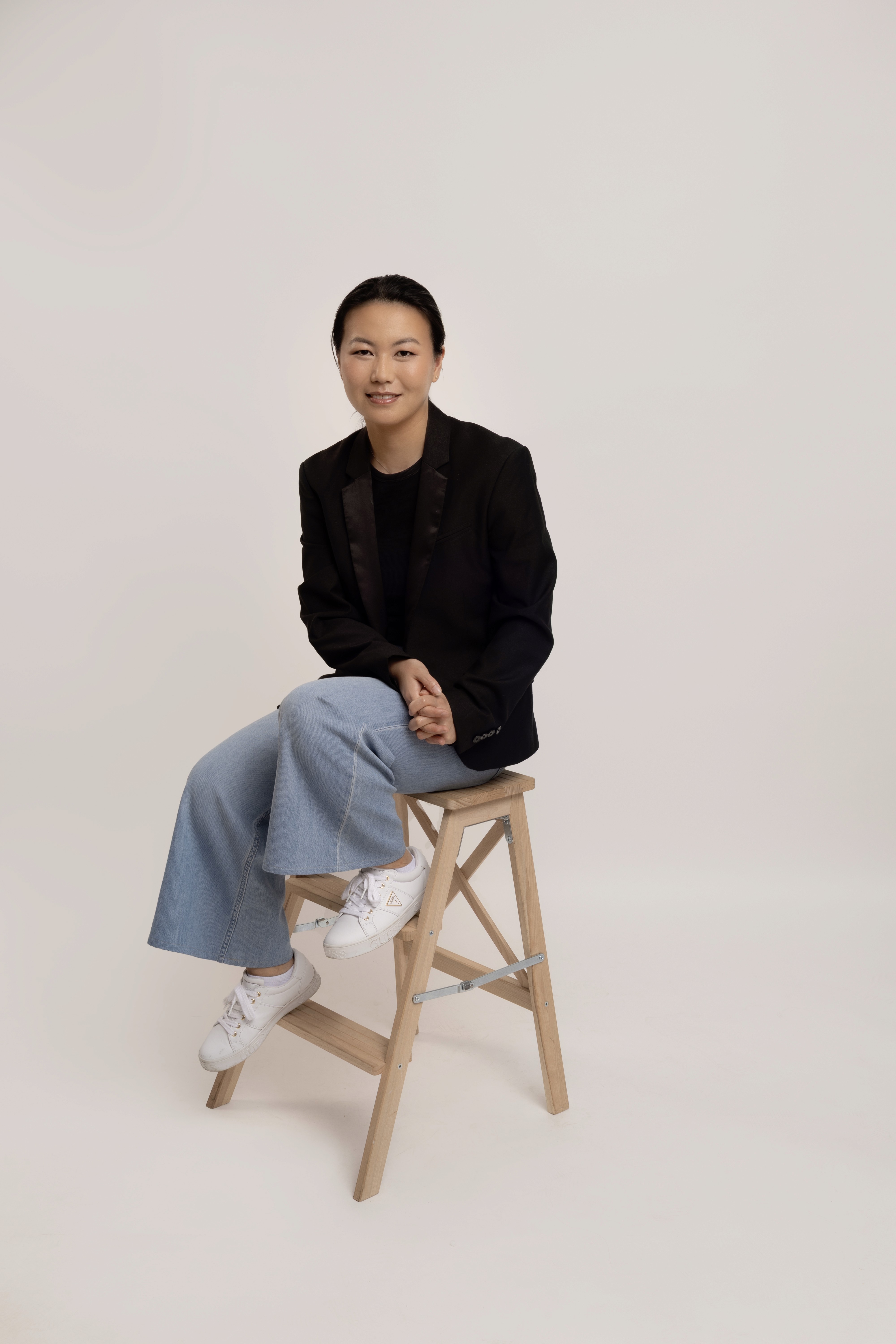 Portrait of Celine sitting on a wooden stool against a soft neutral background, wearing a black blazer, black top, light blue jeans, and white sneakers, with a relaxed and approachable professional expression.
