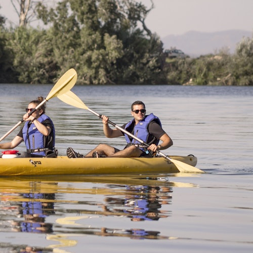 Dos personas con chalecos salvavidas remando en kayak sobre agua tranquila, con árboles y montañas de fondo.