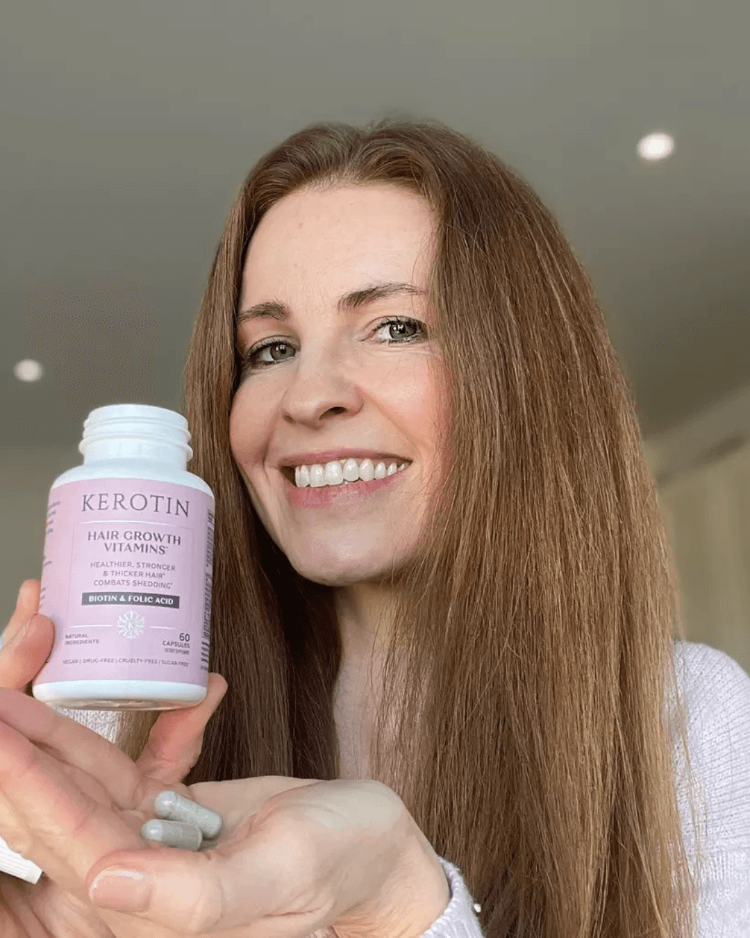 Smiling woman with long hair holds a pink bottle, likely a supplement, against a bright, simple background.
