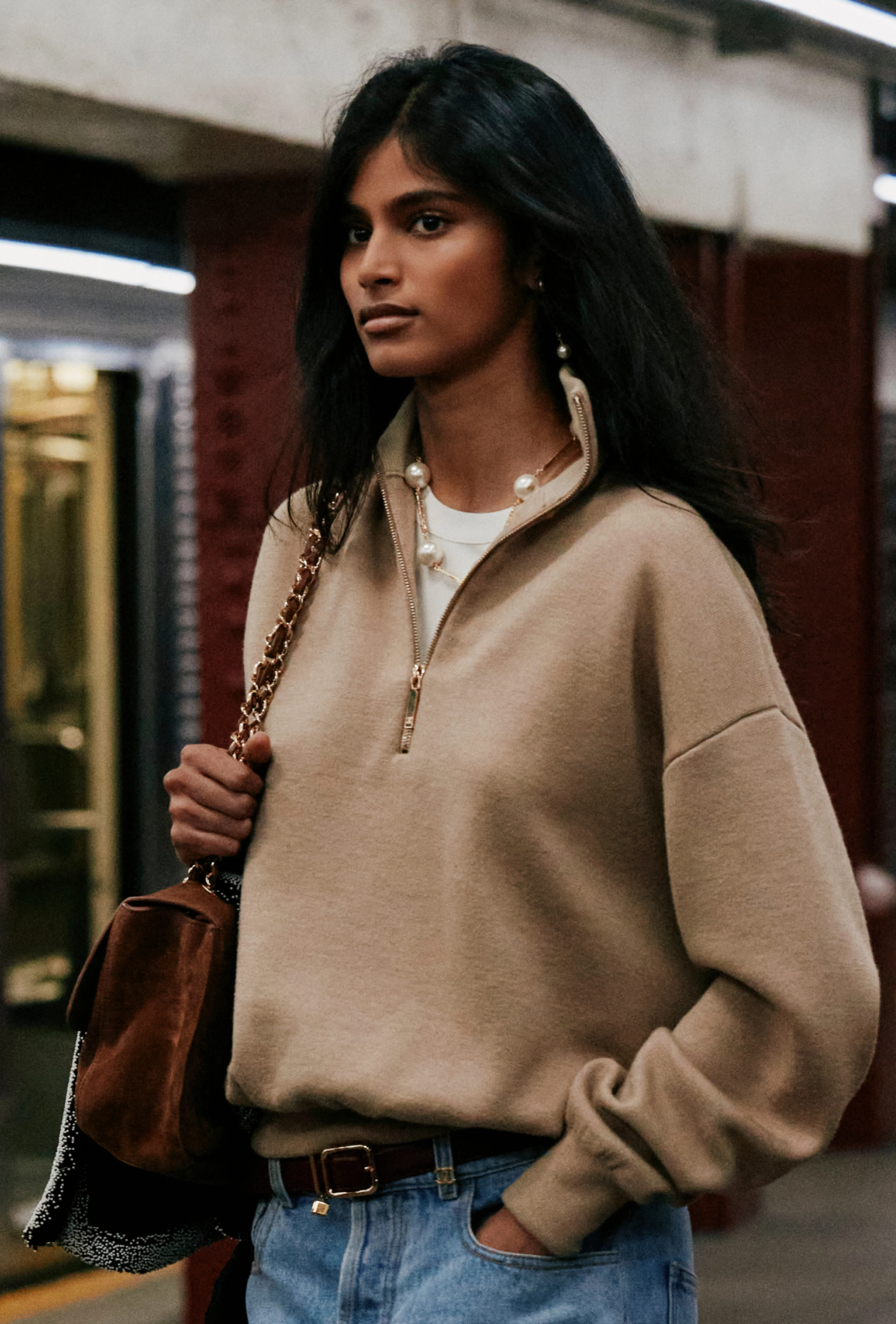 A model with long dark hair wears a beige zip-neck sweater and jeans, carrying a leather bag on a subway platform at the Chanel Métiers d’Art 2026 show.