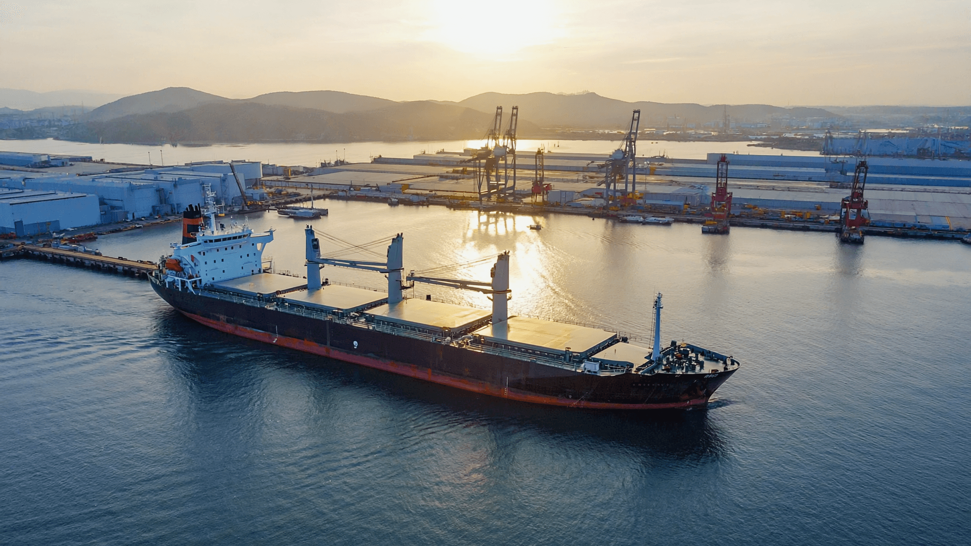 Bulk carrier approaching a Japanese port at sunset, with cranes and terminal facilities in the background.