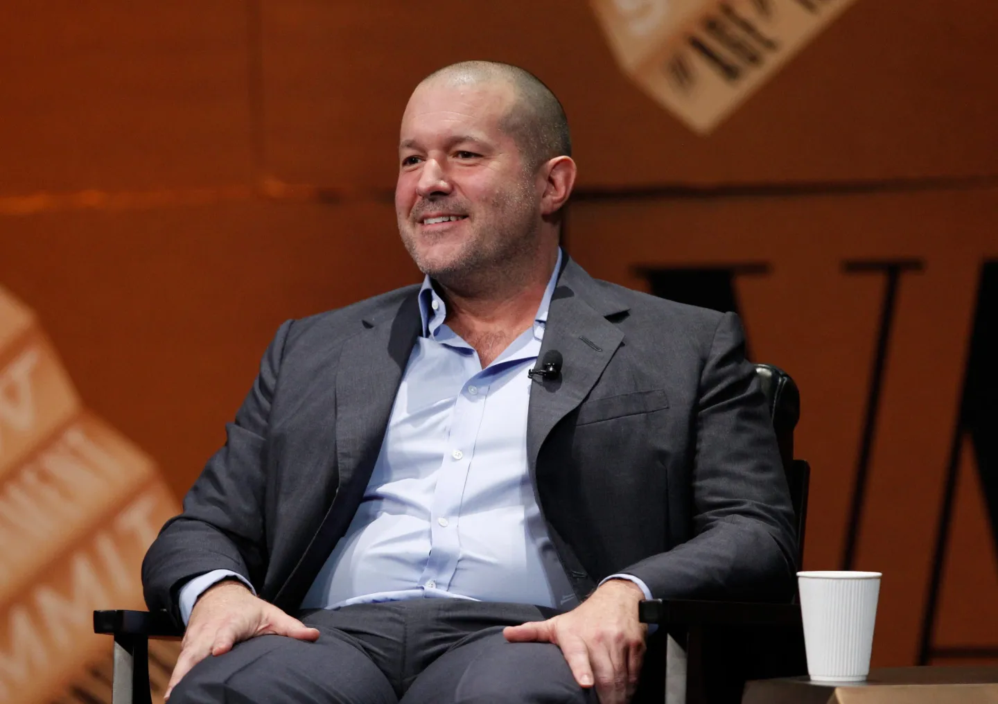 Jony Ive, seated on a chair, wearing a suit and smiling during a talk, highlighting his collaboration with Sam Altman on a new AI hardware project.