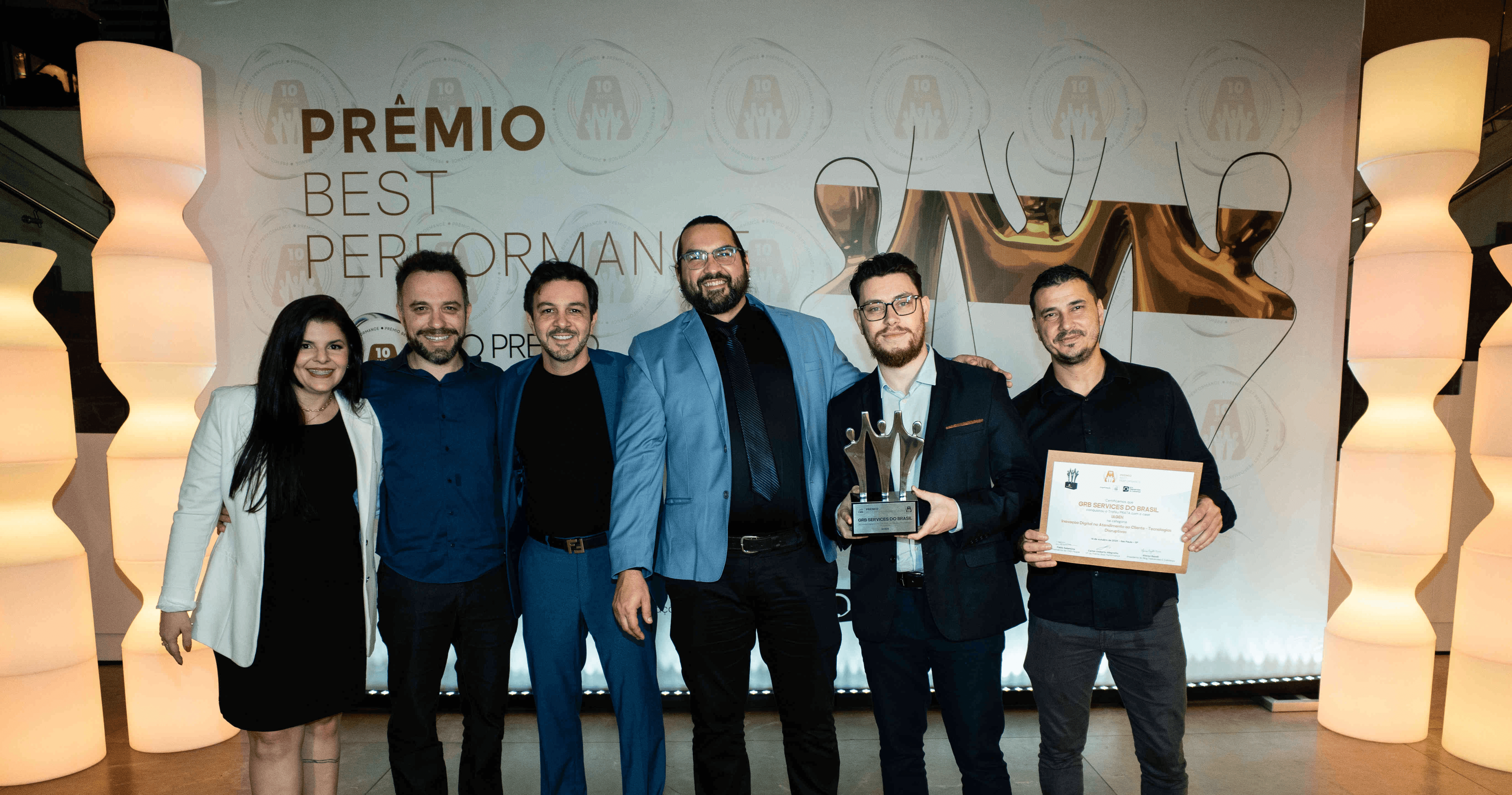 Executivos da GRB SERVICES DO BRASIL posando com troféu prata e certificado no palco do Prêmio Best Performance 2025