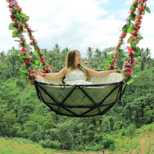 Woman in a floral-adorned swing above a lush, green forest landscape under a partly cloudy sky.