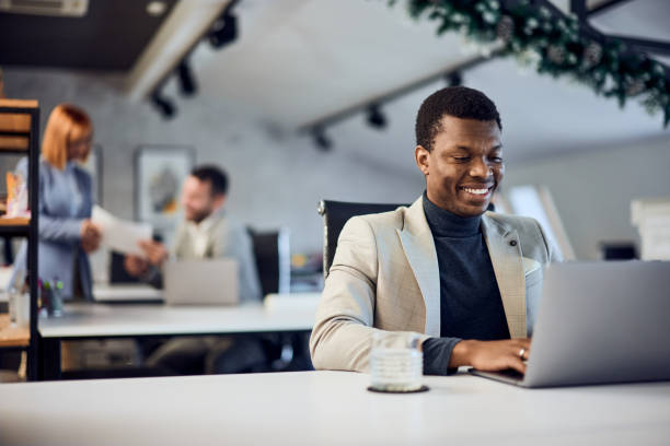 Man smiling while working on a laptop in a modern office, with coworkers collaborating in the background.