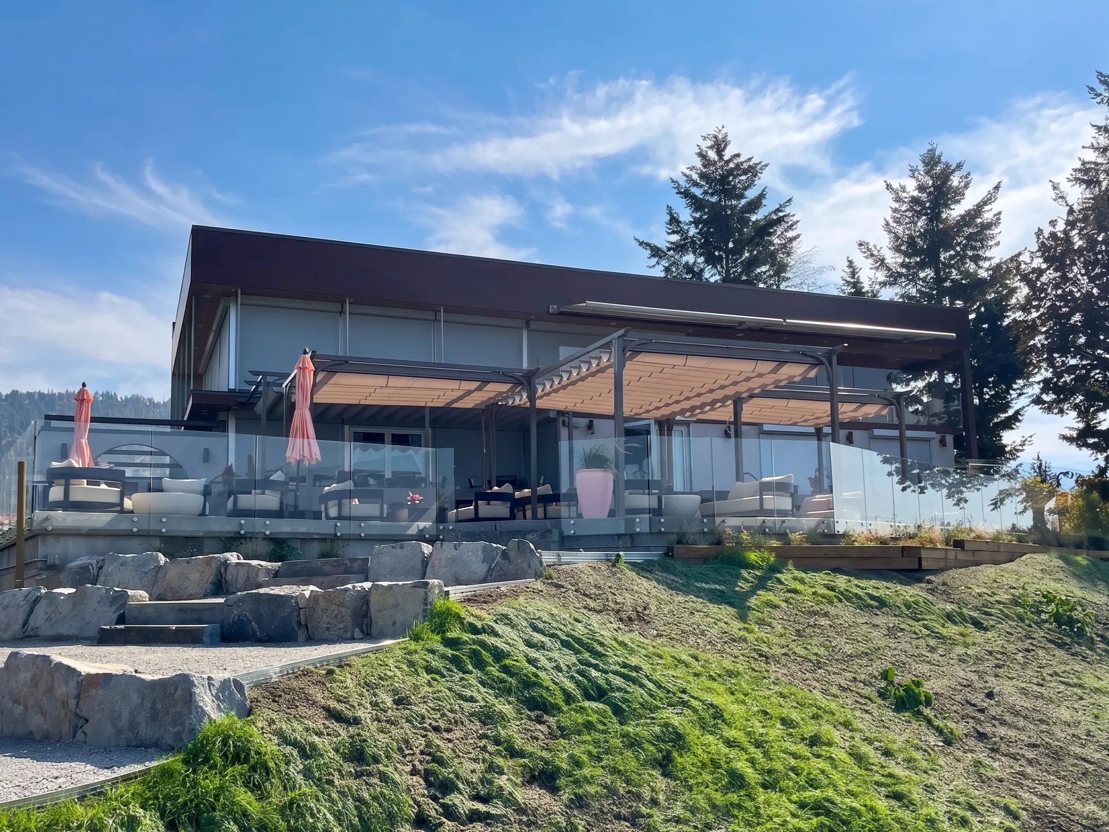 A modern building in Parksville BC with large glass windows and a flat, dark roof is shown on a sunny day. The building has an outdoor patio area with modern, light-colored furniture and a retractable awning or pergola covering part of the space. A glass railing surrounds the patio. In the foreground, a grassy hill slopes up towards the building, with a pathway and large natural rocks forming steps leading up the slope. Tall evergreen trees are visible on the hill behind and to the side of the building.