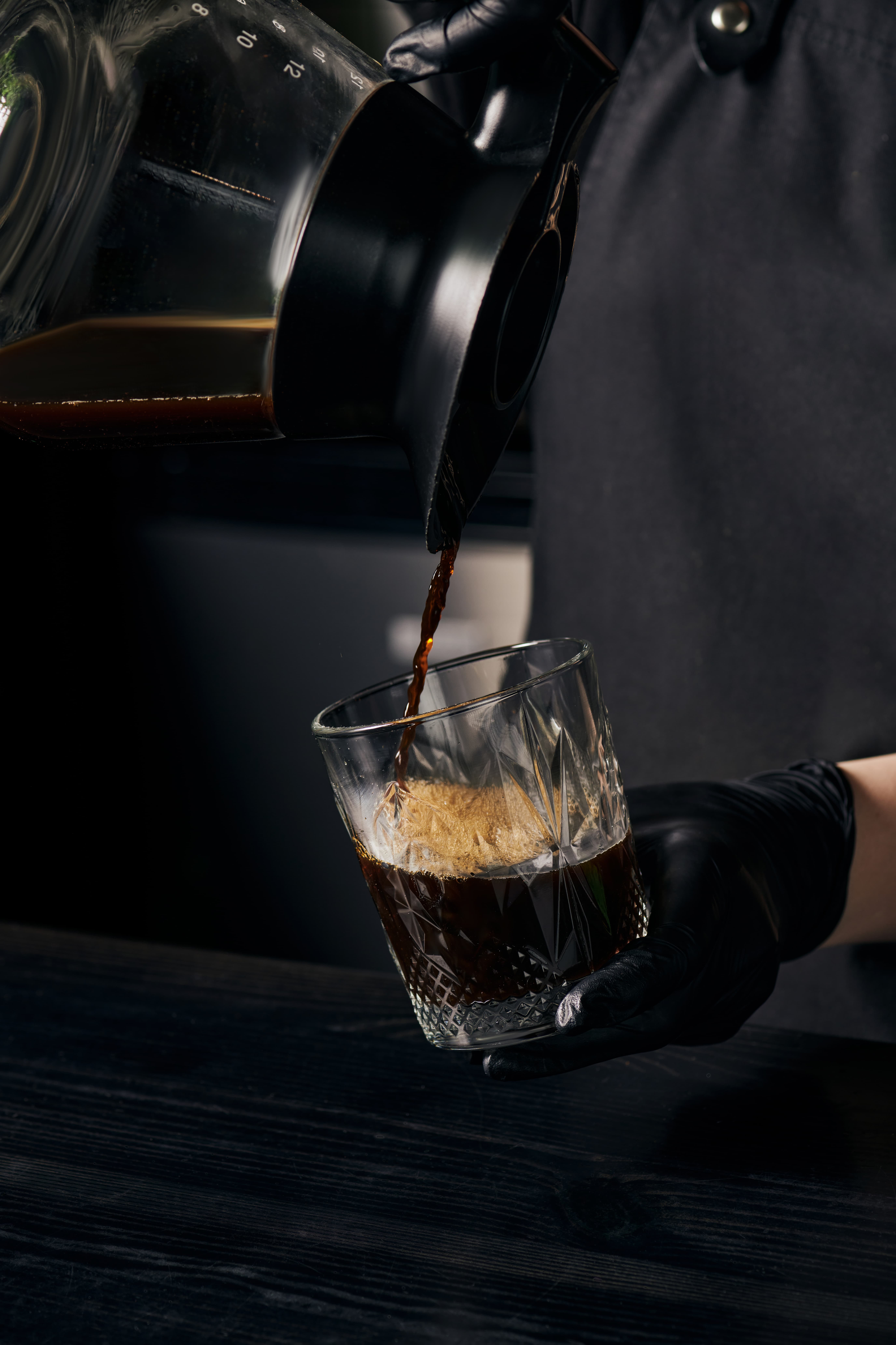 Close-up of coffee being poured from a carafe into a faceted glass, in a dark, moody café setting.