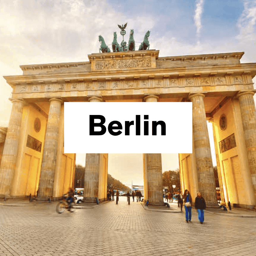 Napis „Berlin” na białej planszy na tle Bramy Brandenburskiej w ciepłym świetle, z przechodniami i rowerzystą