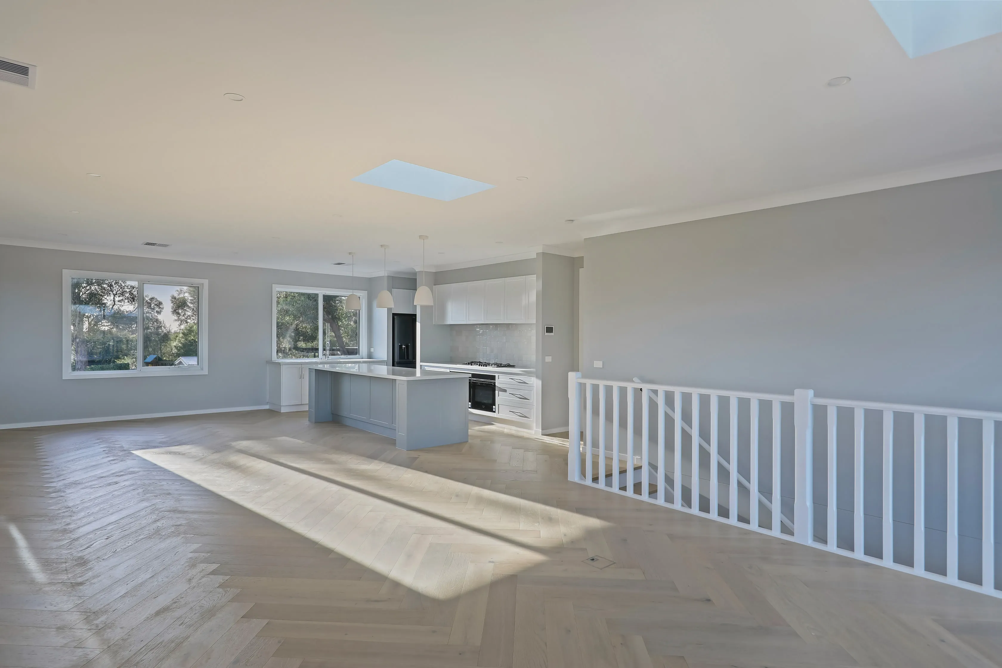 Open living and kitchen with grey island, white pendants, black fridge and oven, dove-grey walls, oak herringbone floors and a white-balustraded stair void with skylights
