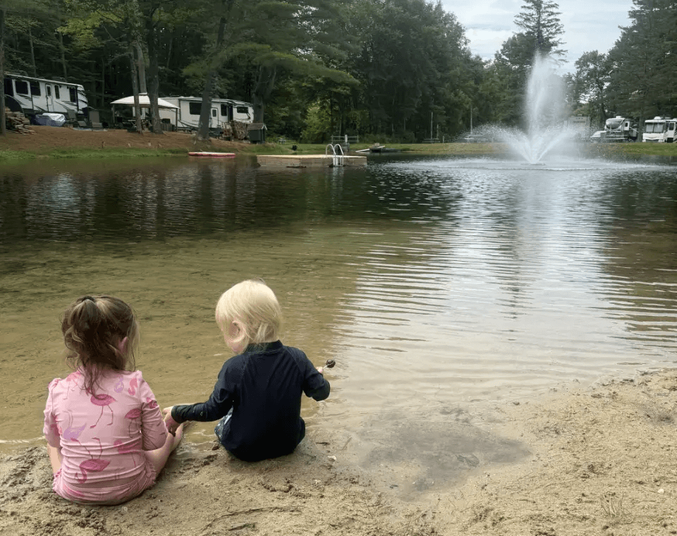 Children sitting on the sandy beach of a private pond with a fountain at Pine Hollow Campground, a top-rated family campground in Southern Vermont near Bennington.