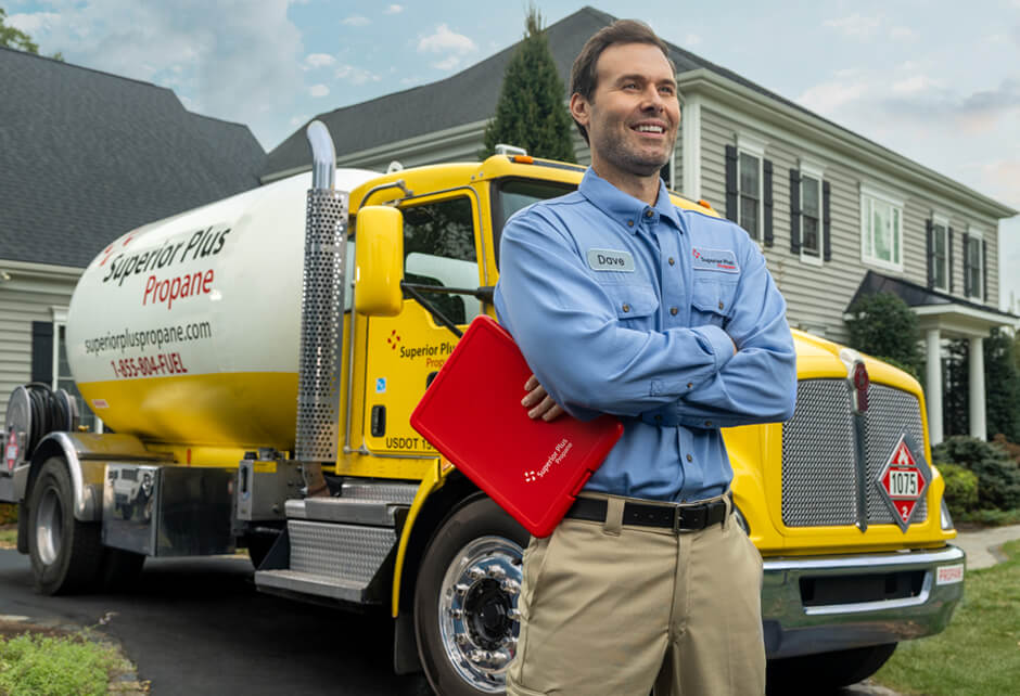 A man in a blue uniform stands in front of a yellow propane delivery truck, holding a red folder, outside a suburban house.