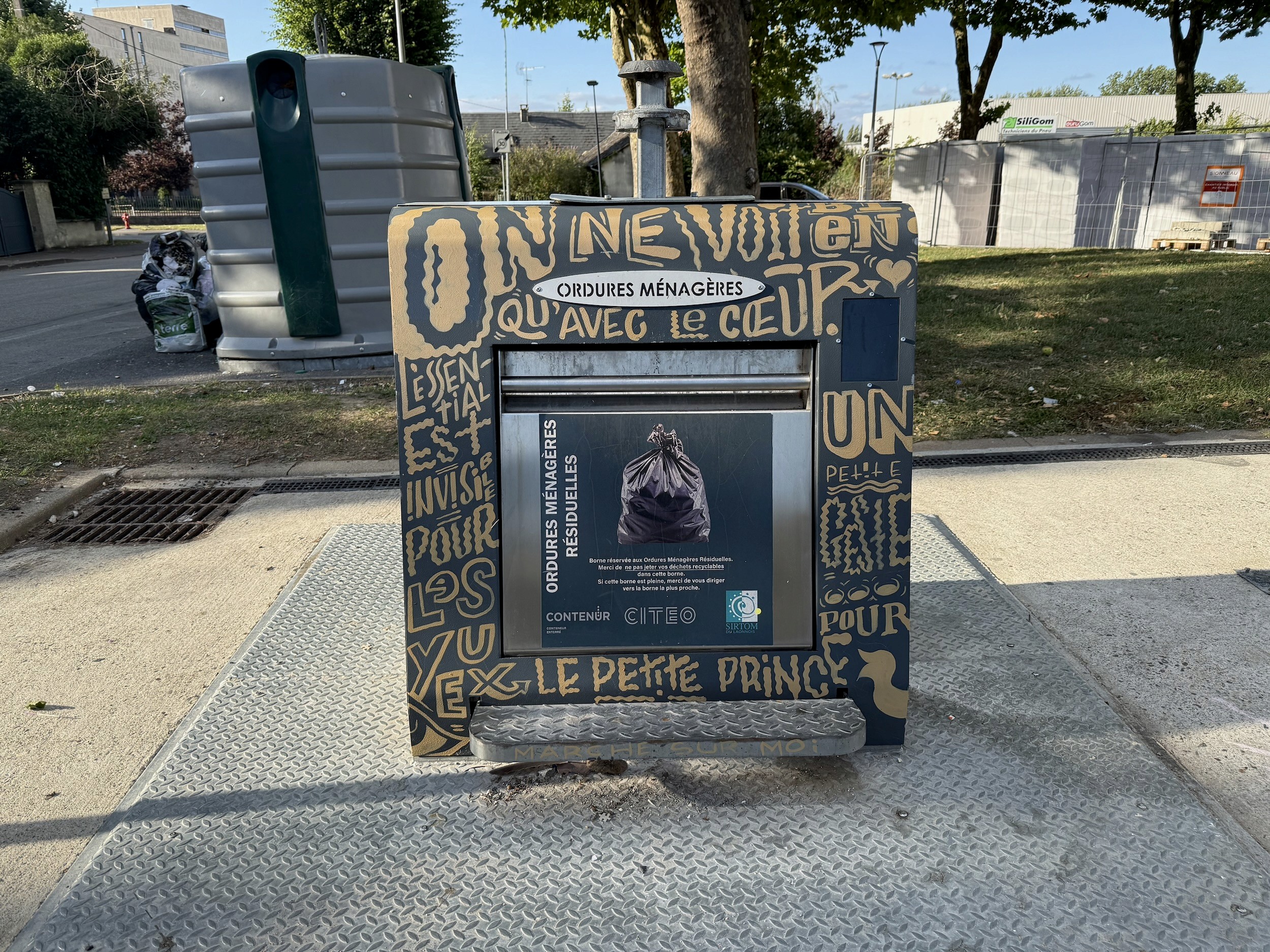 A waste container wrapped in gold hand lettering, “On ne voit bien qu’avec le cœur” running along its top.