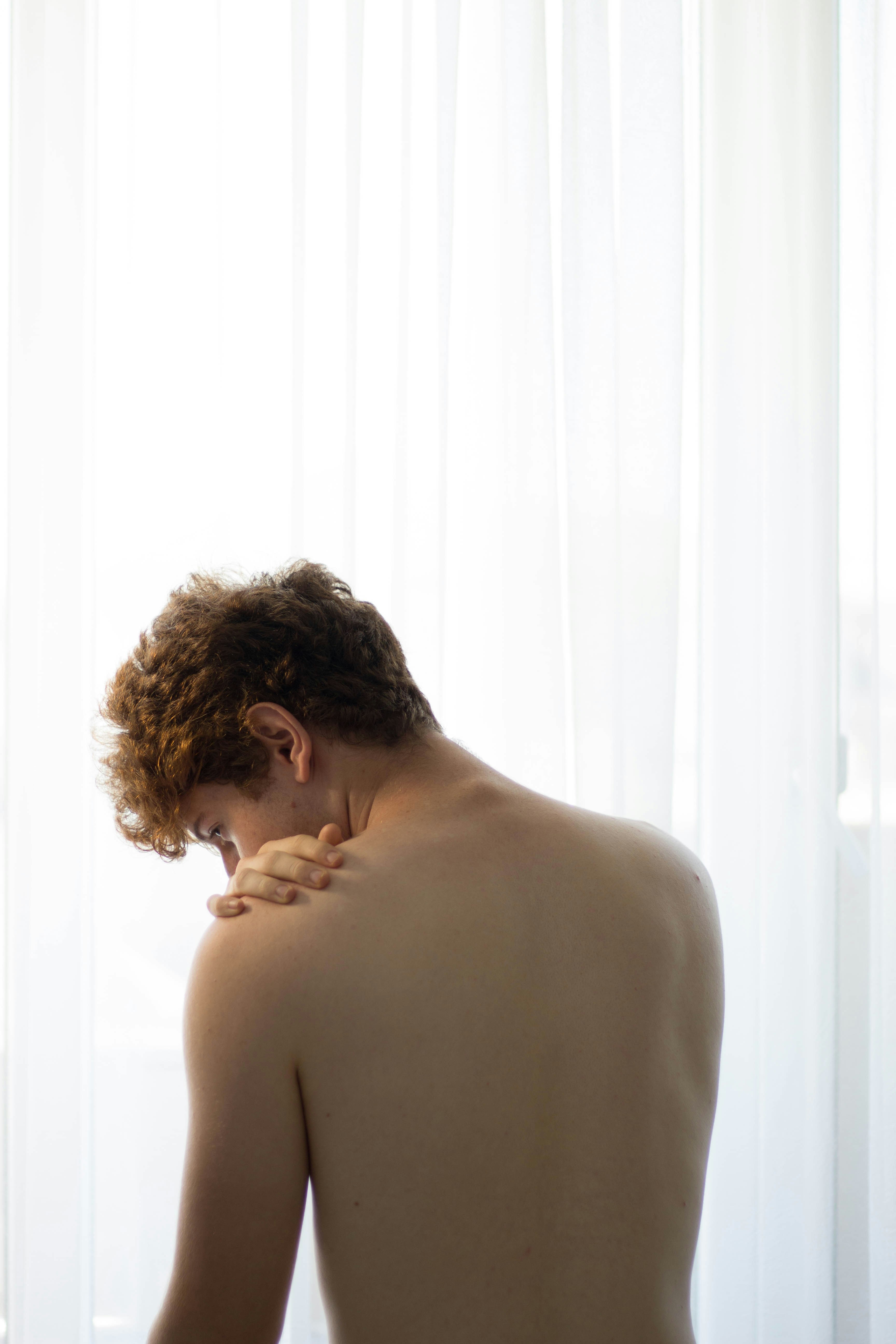 A person with short hair and a bare back is seen from behind, looking down in soft, natural light.
