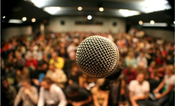 Imagem de um microfone em primeiro plano, com o público ao fundo, desfocado. A foto cria a sensação de estar no palco, prestes a falar para uma audiência ampla e atenta. Ela representa um cenário de palestra ou apresentação pública, capturando a expectativa e a atenção dos espectadores, enquanto o microfone simboliza a comunicação e o compartilhamento de informações em um ambiente profissional ou de entretenimento.