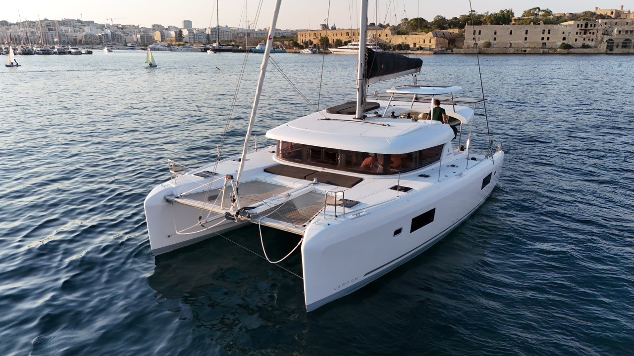 A Lagoon 42 catamaran motoring in a harbor at golden hour, showing its twin hulls, trampoline netting, and flybridge helm station, with a historic waterfront fortress and city skyline behind it.