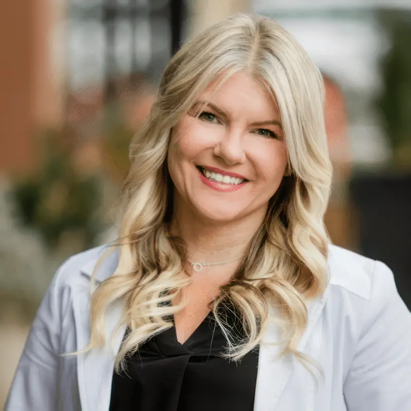 A smiling person with blonde hair, wearing a black top and light-colored blazer, stands outdoors.