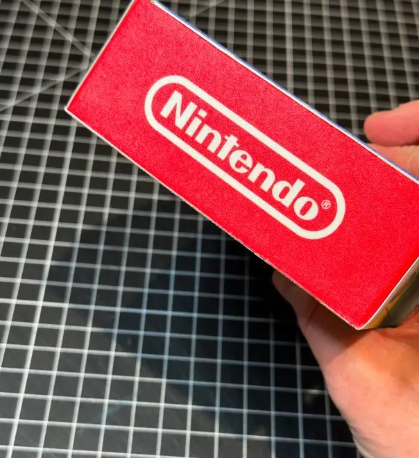 A hand holds a red box showcasing the Nintendo logo, set against a black and white grid surface, highlighting a sleek product design. Rob Harrigan Design. Rob Harrigan. 