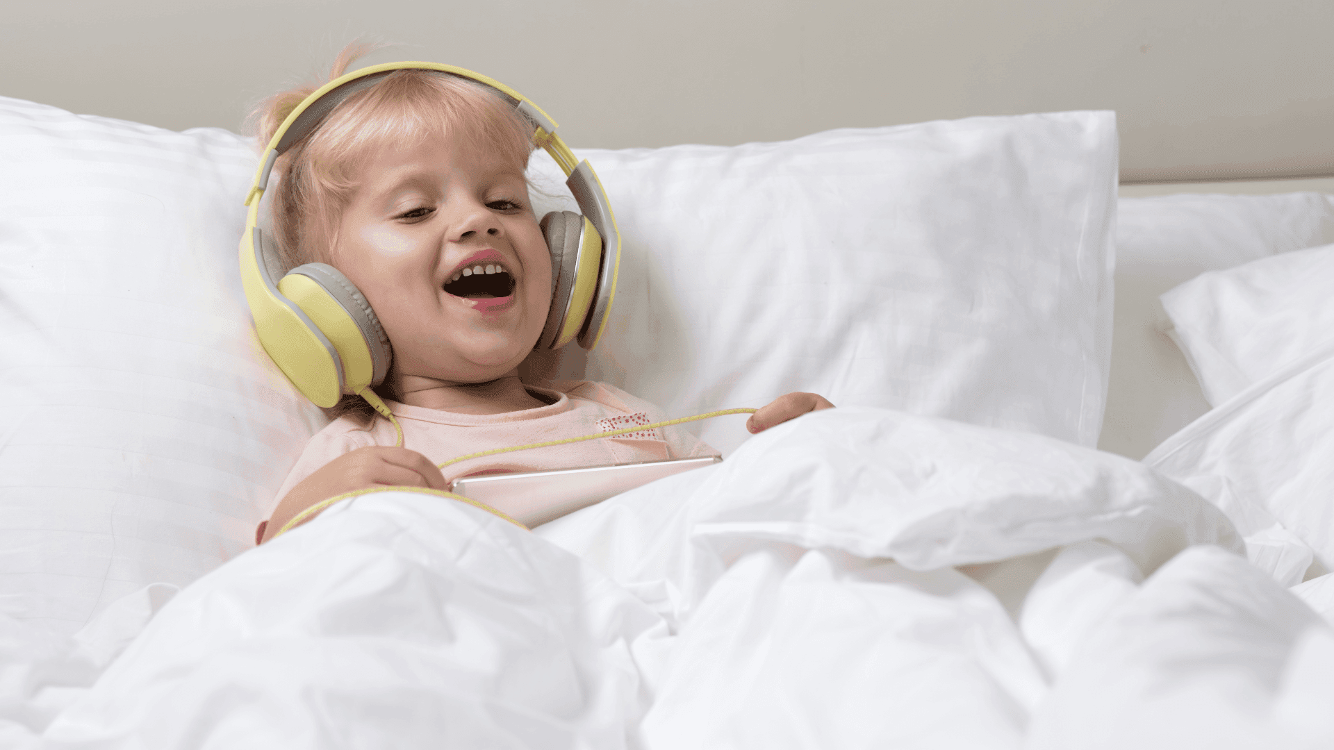 A girl in bed, wearing yellow headphones, smiling and looking happy while listening to HushAway®’s Sound Sanctuary.