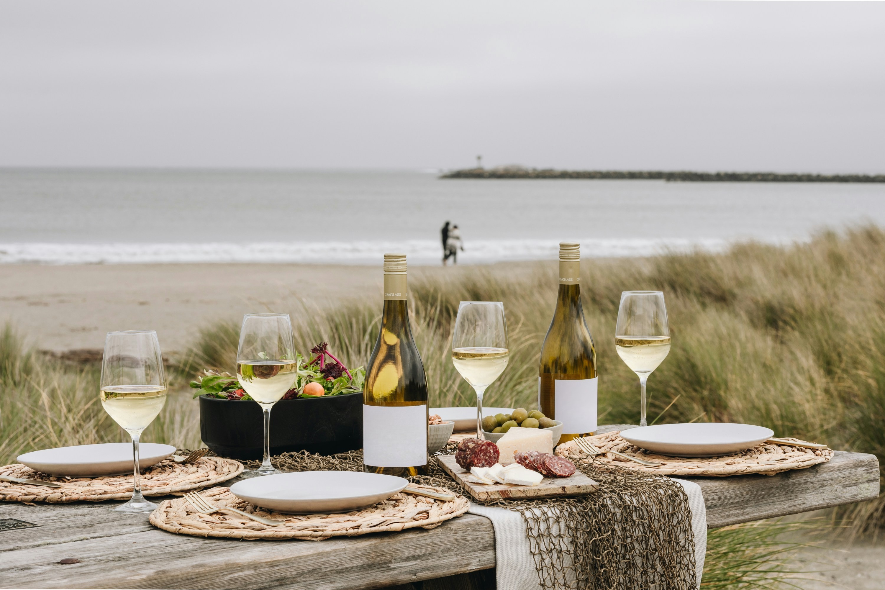 Photo d'un repas posé sur une table avec une salade, de la charcuterie et des bouteilles de vin, dans un paysage naturel et marin