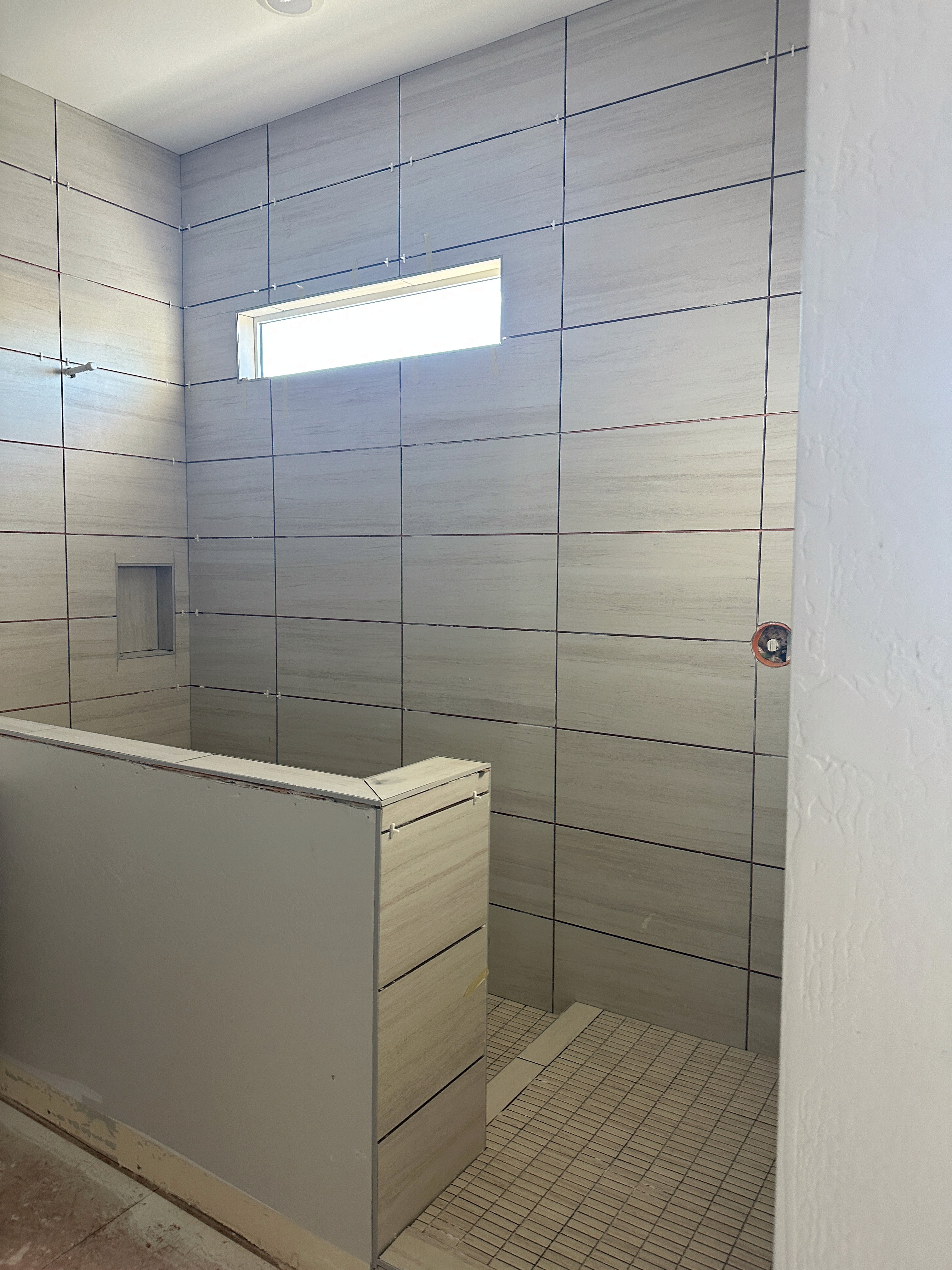 Contemporary shower in progress with horizontal light grey textured wall tiles, a half-wall, a built-in shower niche, and small mosaic floor tiles.