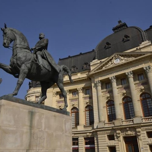 在晴朗的蓝天下，一座宏伟的历史建筑前矗立着一座骑马雕像，该建筑具有圆顶屋顶和柱廊。