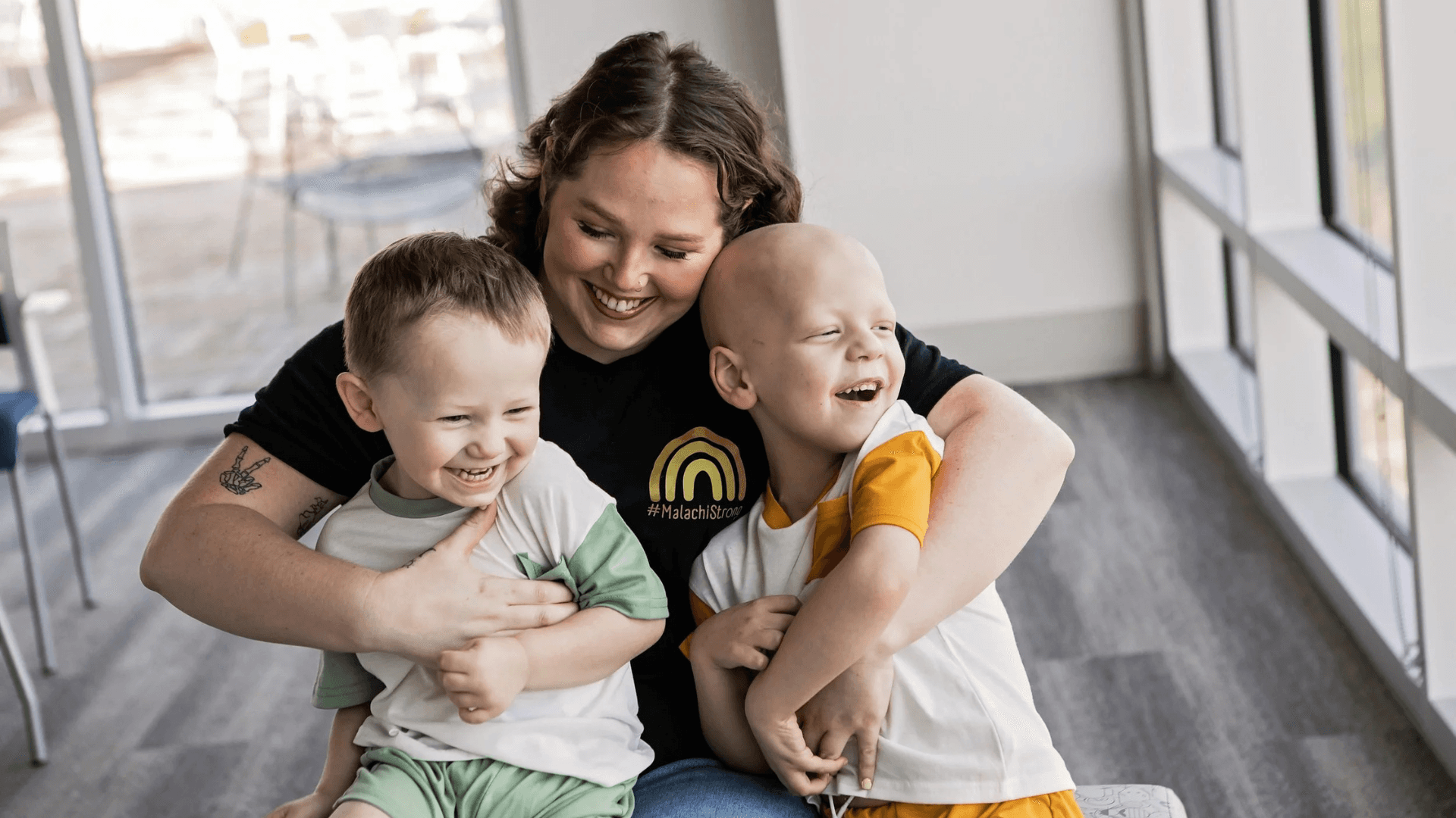 Mother sitting on the floor hugging two young boys, all smiling and laughing together in a bright indoor space with large windows.