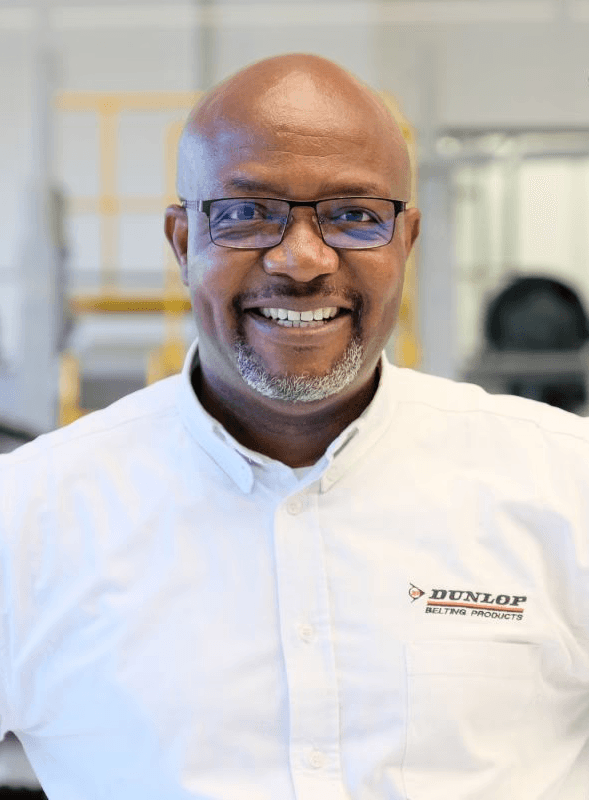 A smiling young man with crossed arms, wearing a plaid shirt and white t-shirt, poses against a dark background.