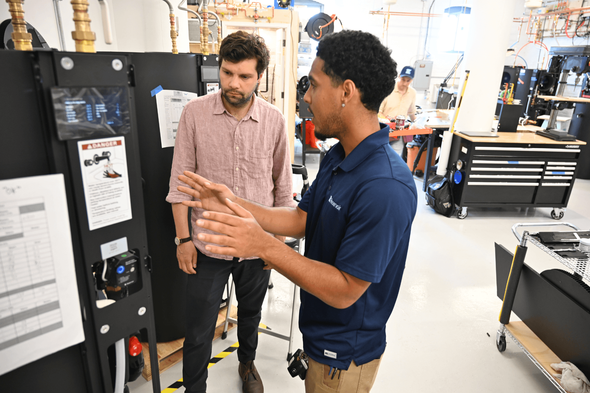 Two men discuss a water heater with its cover off in an engineering workshop.