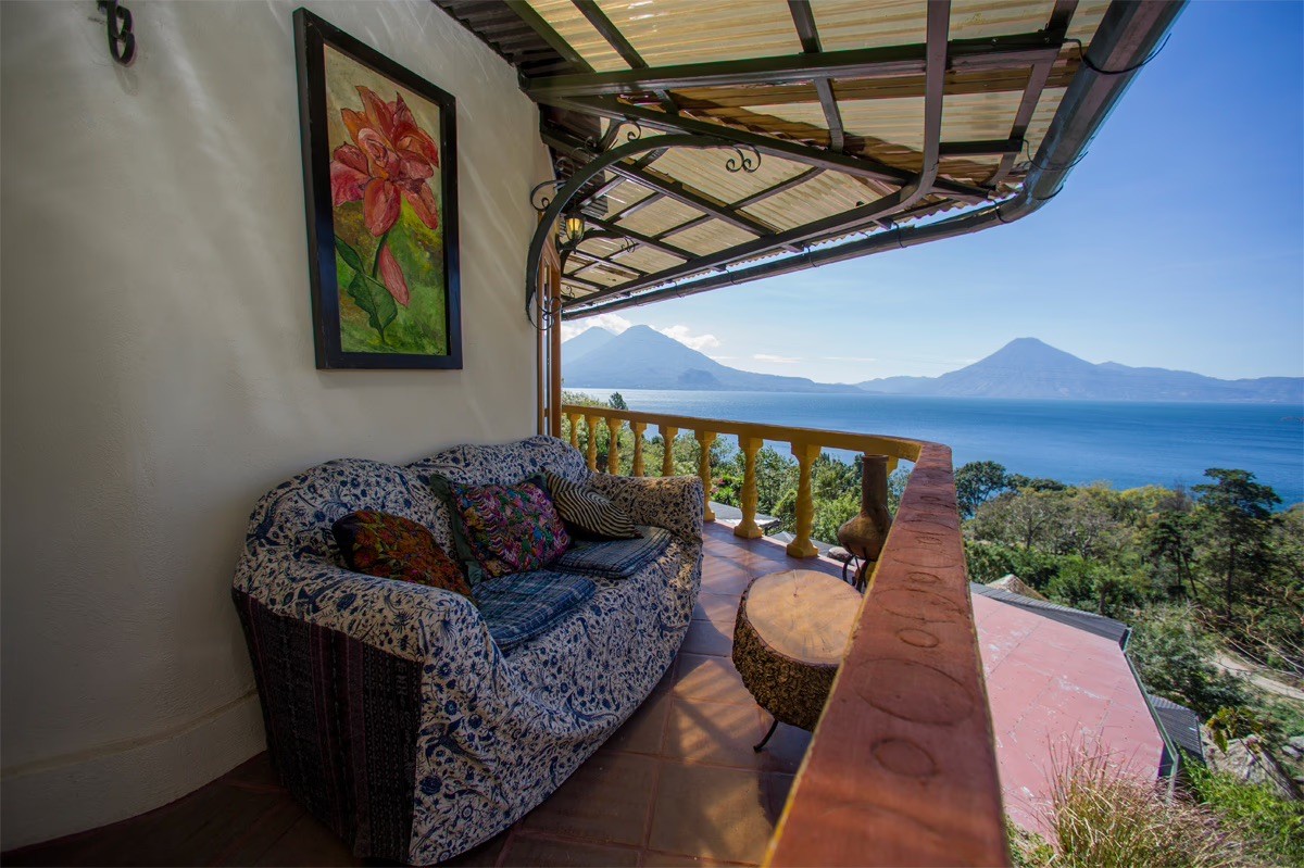 A blue patterned sofa on a balcony with a wood slice table, overlooking a vast blue lake and distant volcanoes.