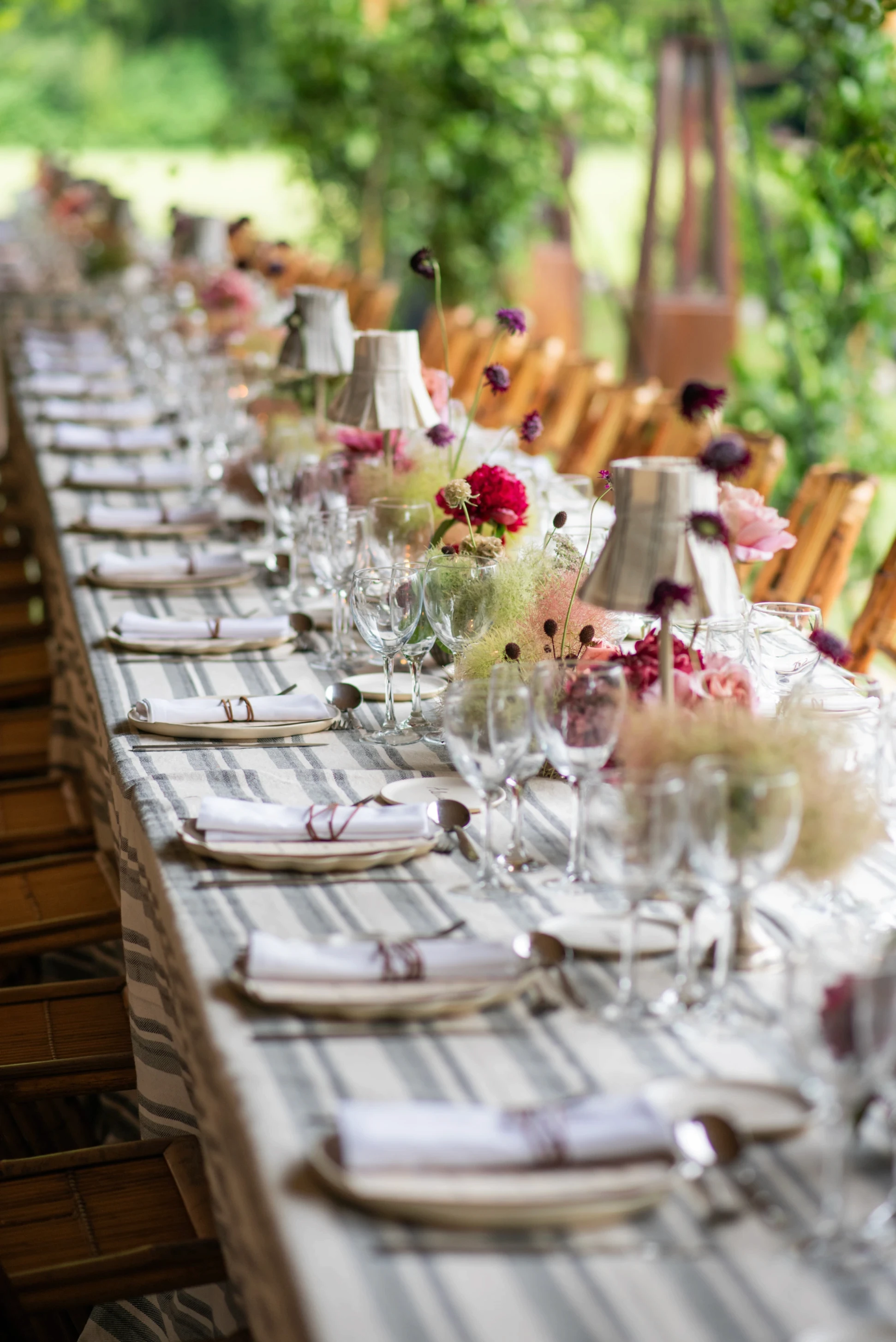 Perspectiva de una mesa larga de boda decorada con gipsofila, smoke bush, flores burdeos y lámparas plisadas.