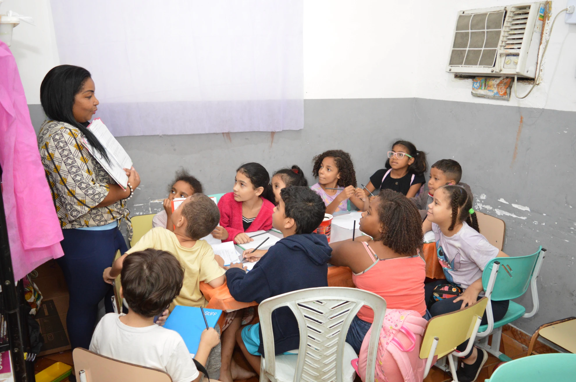 Educadora de pé interagindo com crianças sentadas em sala de aula, todas atentas e envolvidas.