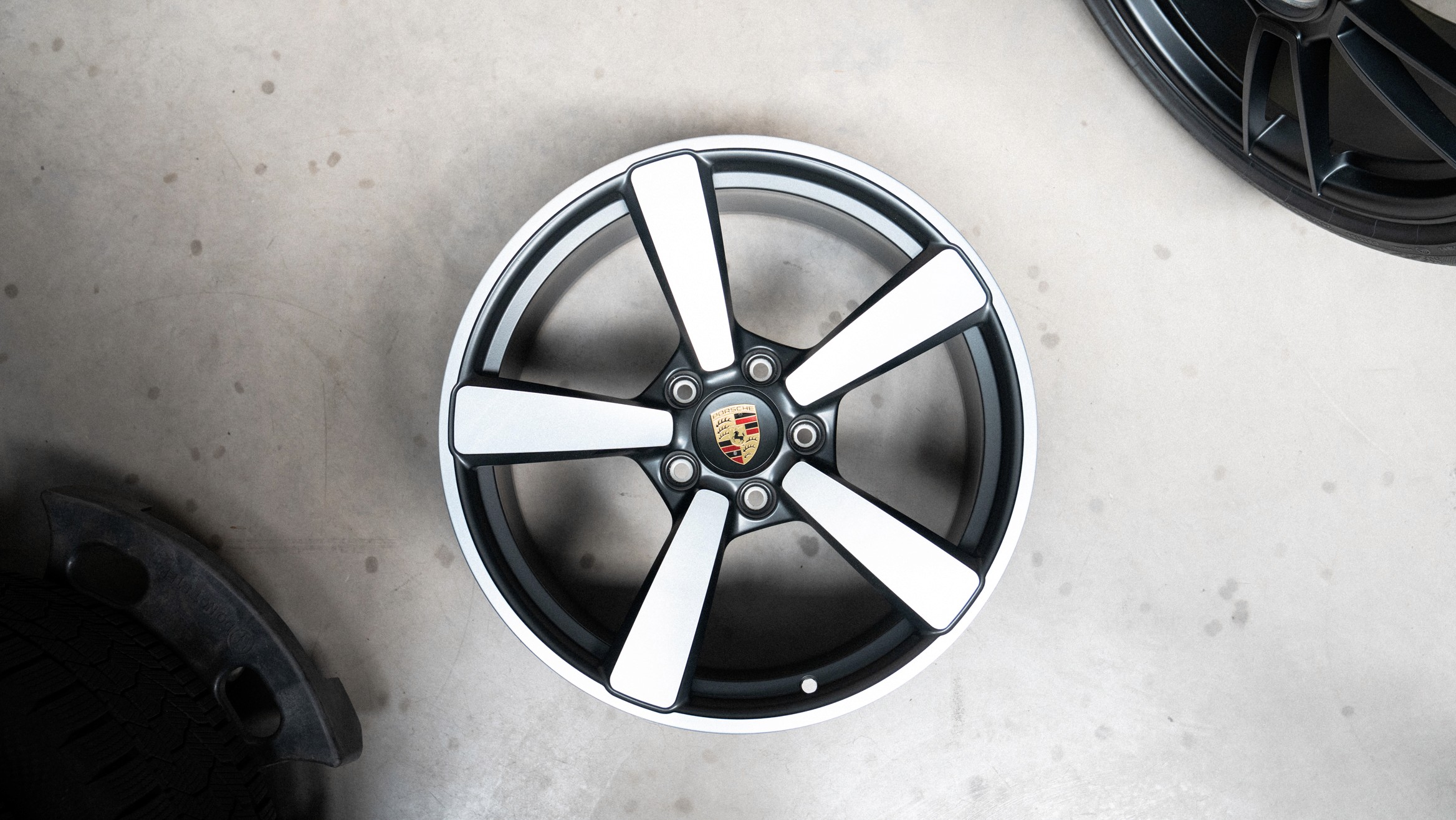 Nahaufnahme einer originalen Porsche-Felge mit fünf Doppelspeichen in Schwarz-Silber und Porsche-Wappen in der Radnabenmitte, auf hellem Untergrund fotografiert.