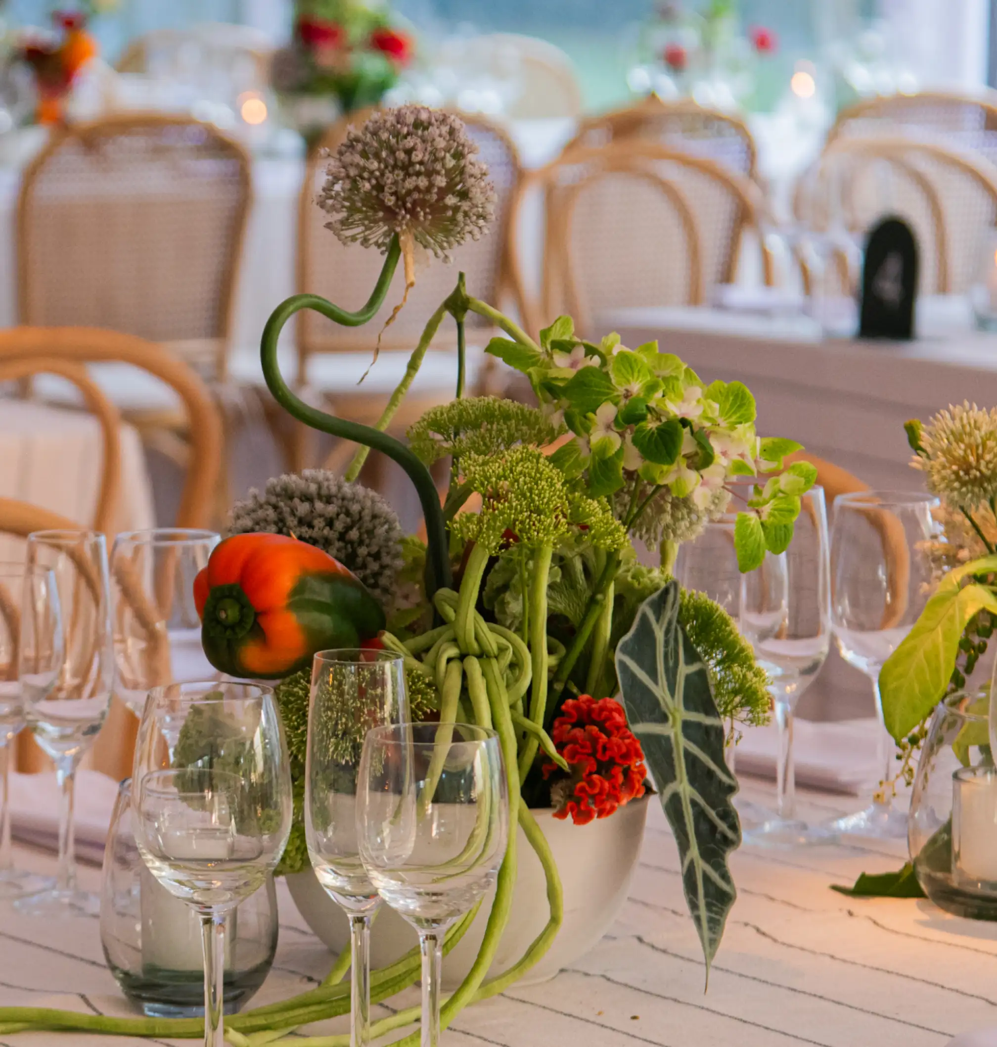 Centro de mesa contemporáneo con flores, follaje y vegetales frescos en tonos verdes, naranjas y rojos, sobre mantel claro y rodeado de copas de cristal.